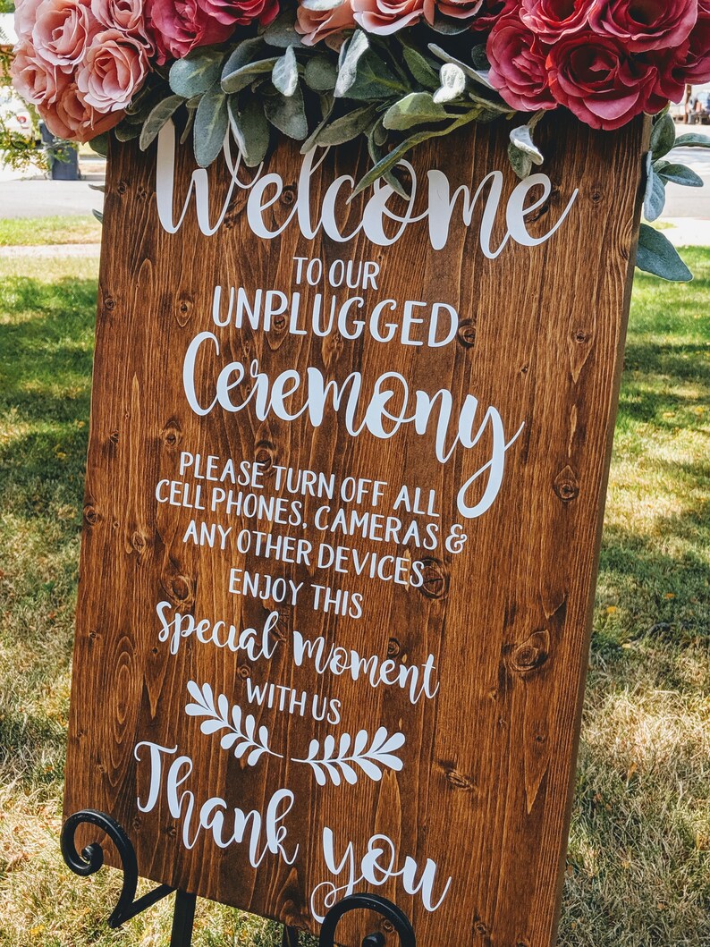 May include: A wooden sign with the text "Welcome to our Unplugged Ceremony. Please turn off all cell phones, cameras & any other devices. Enjoy this special moment with us. Thank you." The sign is decorated with a floral garland.