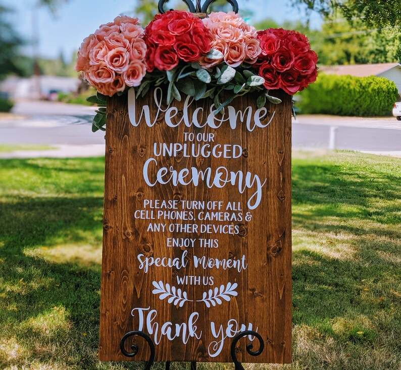 May include: A wooden sign with white text that reads "Welcome to our Unplugged Ceremony. Please turn off all cell phones, cameras & any other devices. Enjoy this special moment with us. Thank you". The sign is decorated with a floral arrangement of pink and red roses.