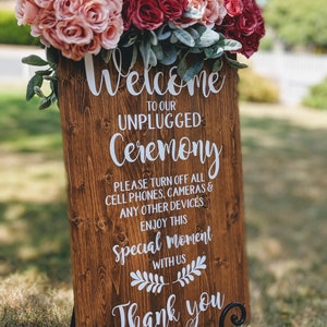 May include: A wooden sign with the text "Welcome to our Unplugged Ceremony" and "Please turn off all cell phones, cameras & any other devices. Enjoy this special moment with us. Thank you." The sign is decorated with pink and red flowers.