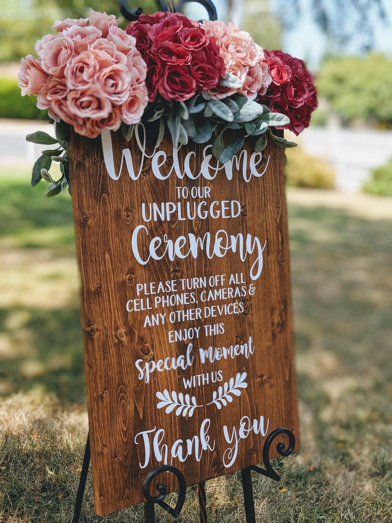 May include: A wooden sign with the text "Welcome to our Unplugged Ceremony" and "Please turn off all cell phones, cameras & any other devices. Enjoy this special moment with us. Thank you." The sign is decorated with pink and red flowers.
