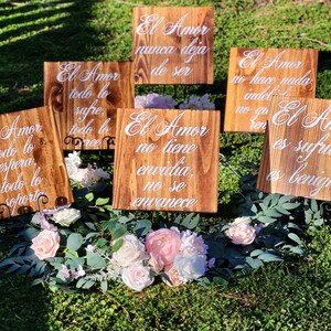 May include: Six wooden signs with white text on a brown background. The signs are arranged in a circle on a bed of green grass and pink flowers. The text on the signs reads: "El Amor nunca deja de ser", "El Amor todo lo sufre, todo lo cree", "El Amor todo lo espera, todo lo soporta", "El Amor no tiene envidia, no se envanece", "El Amor no hace nada indebido, no quiere ren", "El Amor es sufrido, es benigno".