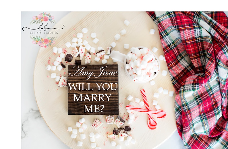 May include: A wooden sign with the text "Amy Jane Will You Marry Me?" surrounded by white marshmallows, candy canes, and chocolate pieces on a wooden surface.
