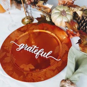 May include: A close-up of a round, orange charger plate with a leaf design. The word "grateful" is written in white on the plate.