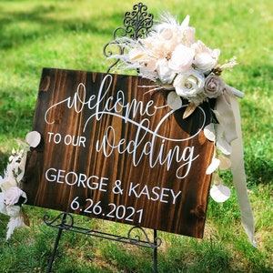 May include: A wooden wedding sign with white calligraphy that reads "Welcome to our wedding George & Kasey 6.26.2021". The sign is decorated with a bouquet of white and blush flowers and greenery.
