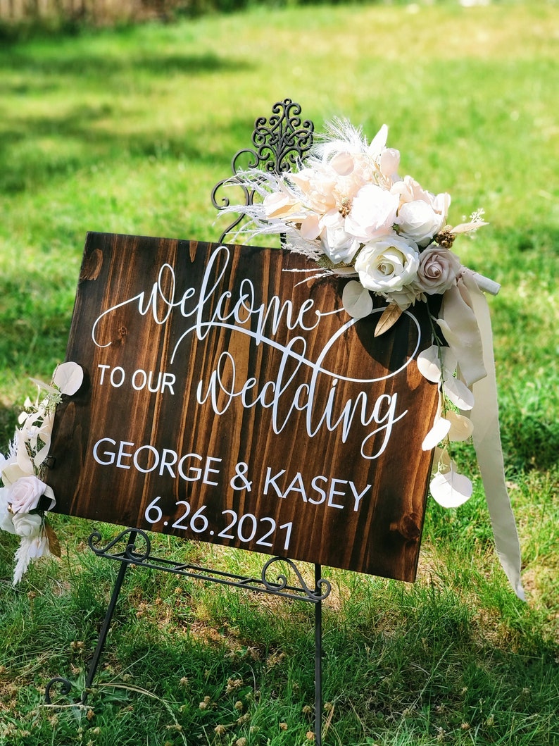 May include: A wooden wedding sign with white calligraphy that reads "Welcome to our wedding George & Kasey 6.26.2021". The sign is decorated with a bouquet of white and blush flowers and greenery.