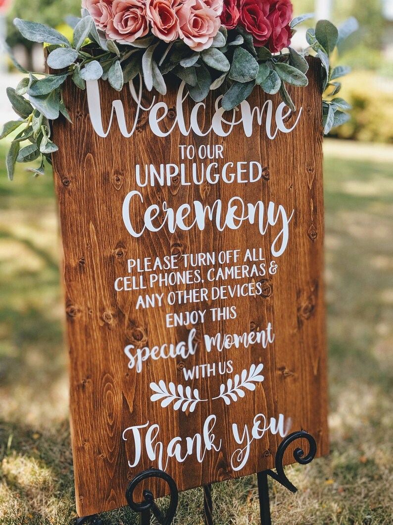May include: A wooden sign with white text that reads "Welcome to our Unplugged Ceremony. Please turn off all cell phones, cameras & any other devices. Enjoy this special moment with us. Thank you". The sign is decorated with a floral arrangement of pink and red roses and greenery.