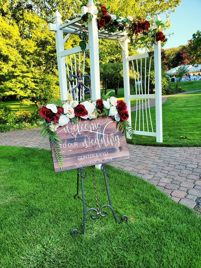 May include: A wooden sign with the text "Welcome to our Wedding" and the names "Gunter & Kyli" and the date "September 14, 2019". The sign is on an easel in front of a white wooden archway decorated with red and white flowers.