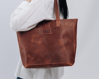 May include: A woman wearing a white shirt and blue jeans is holding a brown leather tote bag with a small pocket on the front. The bag has a leather strap and a small logo on the front.