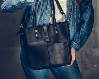May include: A black leather tote bag with a top handle and a shoulder strap. The bag has a large front pocket and a smaller pocket on the side.