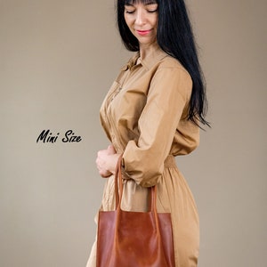 Pu&ograve; includere: Una donna indossa una tuta beige e tiene una borsa tote in pelle marrone. La borsa ha un design semplice con due manici. Il testo "Mini Size" &egrave; scritto in corsivo nero sul lato sinistro dell'immagine.