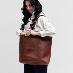 May include: A large, rectangular brown leather tote bag with two shoulder straps. The bag features a smooth, matte finish with visible stitching at the base of the handles. The design is simple and functional, providing a spacious interior compartment.