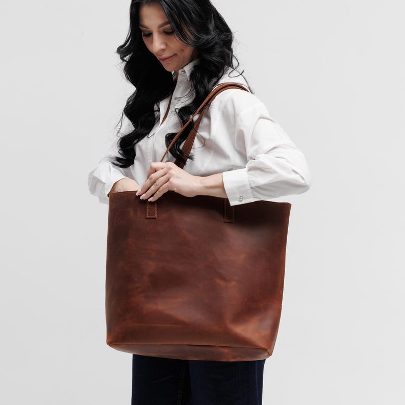May include: A large, rectangular brown leather tote bag with two shoulder straps. The bag features a smooth, matte finish with visible stitching at the base of the handles. The design is simple and functional, providing a spacious interior compartment.
