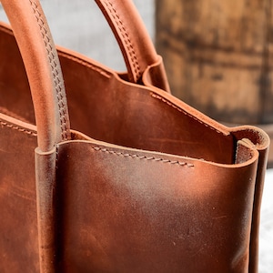 May include: Close-up of a brown leather tote bag with a stitched handle. The bag is made of high-quality leather and has a classic design.