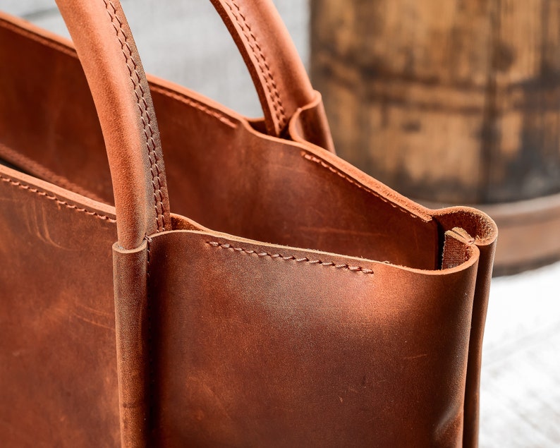 May include: Close-up of a brown leather tote bag with a stitched handle. The bag is made of high-quality leather and has a classic design.
