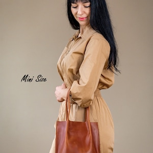 May include: A woman wearing a tan jumpsuit holds a brown leather tote bag. The bag has a simple design with two handles. The text "Mini Size" is written in black cursive on the left side of the image.