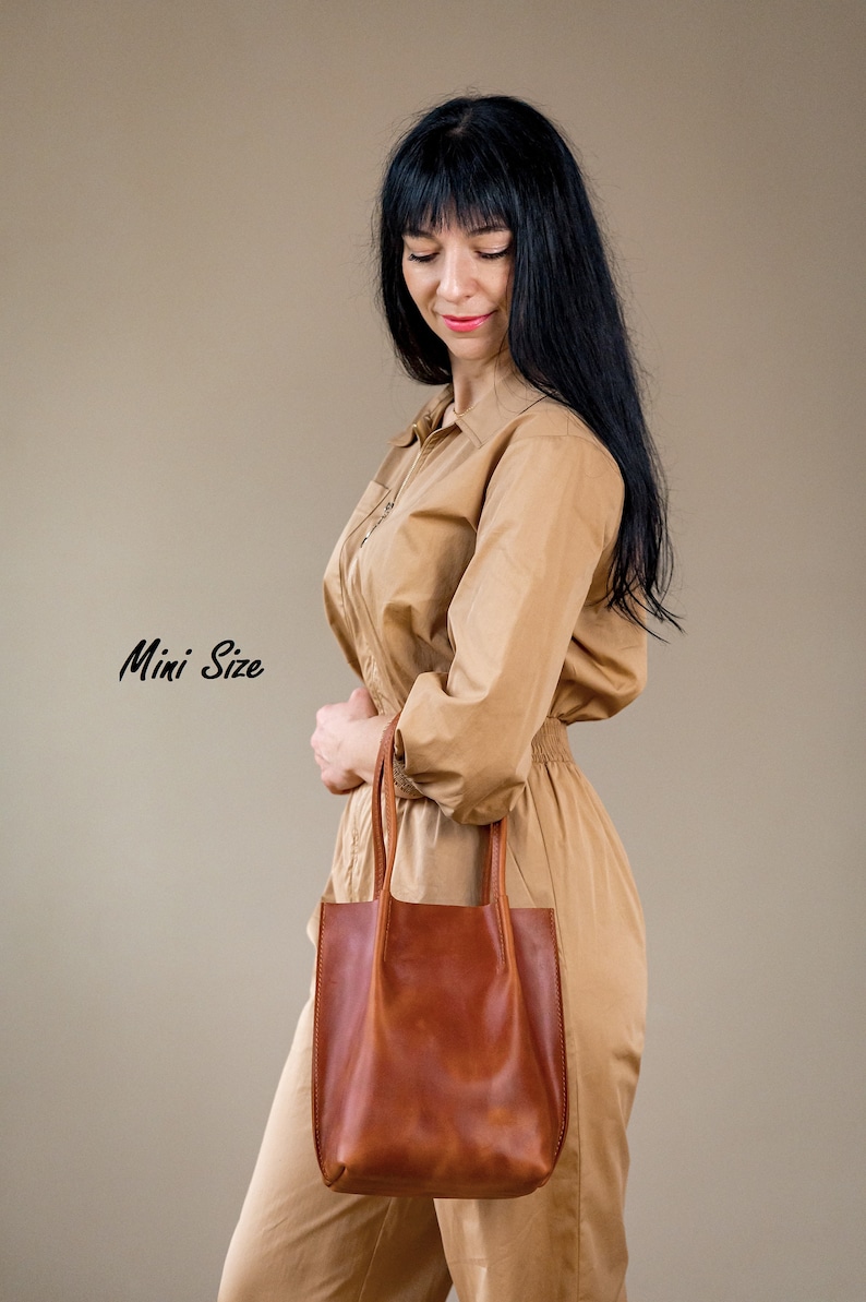 May include: A woman wearing a tan jumpsuit holds a brown leather tote bag. The bag has a simple design with two handles. The text "Mini Size" is written in black cursive on the left side of the image.