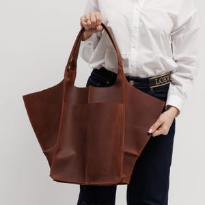 Pu&ograve; includere: Una grande borsa tote strutturata in pelle marrone con una silhouette ampia e svasata e due manici. La borsa presenta una cucitura verticale centrale, impunture visibili lungo i bordi e un design spazioso a giorno con una finitura opaca.