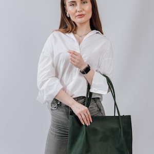 May include: A woman wearing a white button-down shirt and gray jeans is holding a dark green leather tote bag.