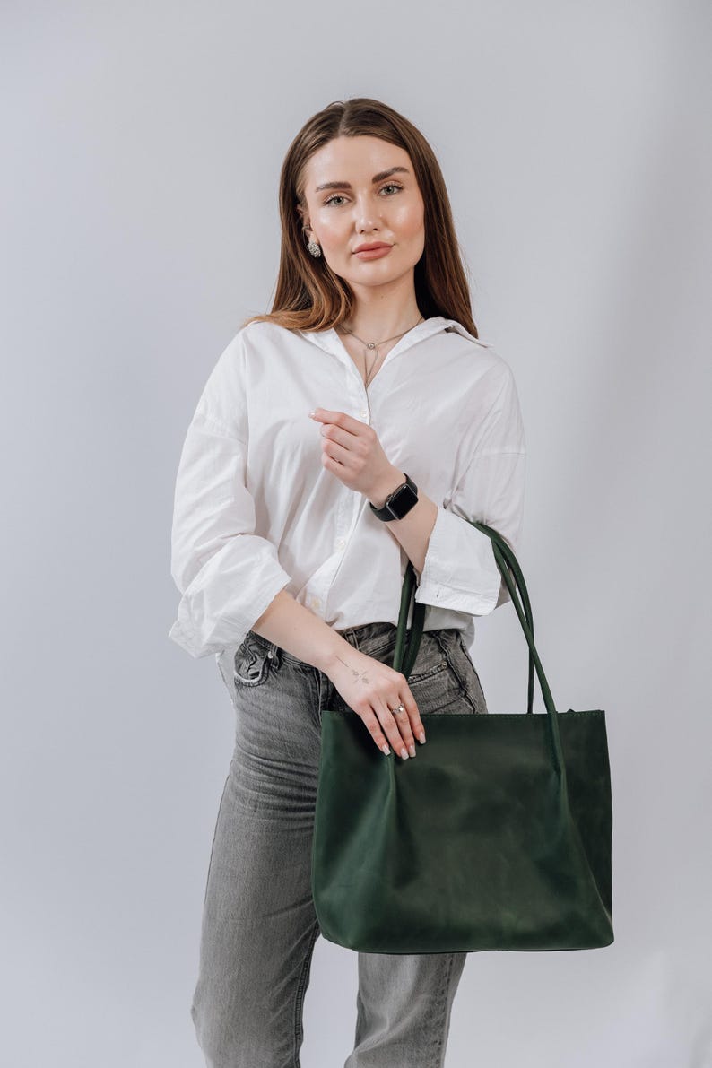 May include: A woman wearing a white button-down shirt and gray jeans is holding a dark green leather tote bag.