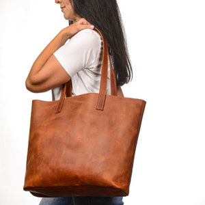 Op de afbeelding: Een bruine leren tote bag met een brede opening en twee lange hengsels. De tas wordt gedragen door iemand die een wit T-shirt en een blauwe jeans draagt.