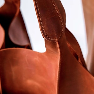 Pu&ograve; includere: Una borsa tote in pelle marrone medio con spallacci in pelle spessi e robusti, fissati saldamente con cuciture visibili. La borsa presenta una finitura liscia e opaca e un design strutturato a giorno con pannelli laterali profondi e curvi.