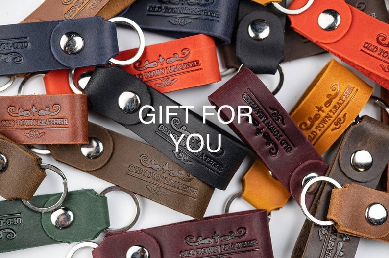 Op de afbeelding: Assortiment van leren sleutelhangers met een zilveren sleutelring en een metalen knoop. Elke sleutelhanger heeft de tekst "Old Town Leather" erop gegraveerd. De tekst "Gift for You" is over de afbeelding gelegd.
