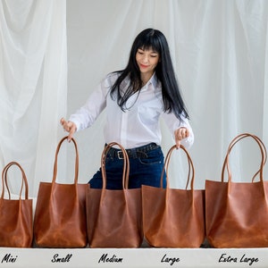 May include: Five brown leather tote bags of different sizes lined up on a white surface. The bags are labeled "Mini", "Small", "Medium", "Large", and "Extra Large".