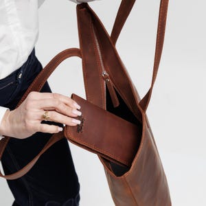 May include: A brown leather tote bag with thin shoulder straps and a silver-toned metal zipper closure. A matching brown leather wallet with a small embossed logo is being placed into the open main compartment of the bag.