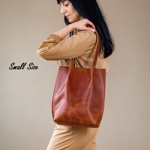 May include: A woman is holding a brown leather tote bag. The bag has a simple design and is made of high-quality leather. The text "Small Size" is visible in the image.
