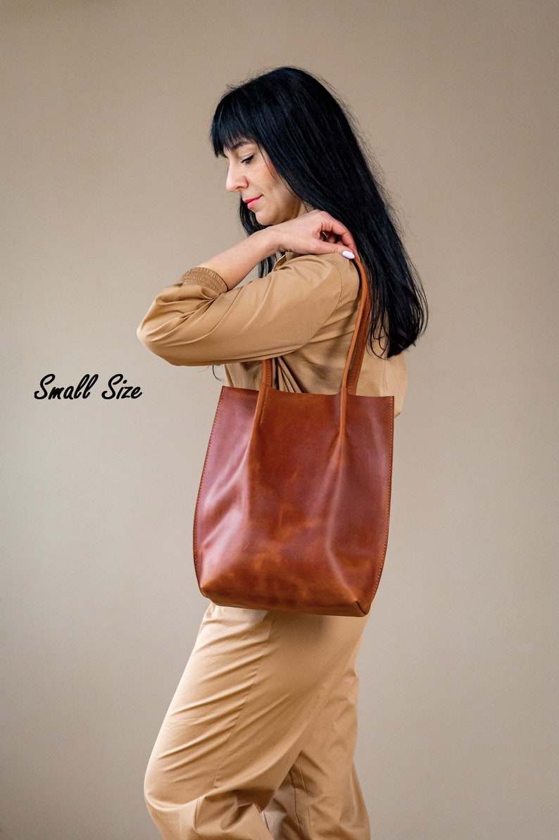 May include: A woman is holding a brown leather tote bag. The bag has a simple design and is made of high-quality leather. The text "Small Size" is visible in the image.