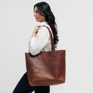 May include: A large, rectangular tote bag crafted from smooth, dark brown leather. The bag features two long, sturdy shoulder straps securely stitched to the top edge, providing a clean, minimalist silhouette with a spacious main compartment.