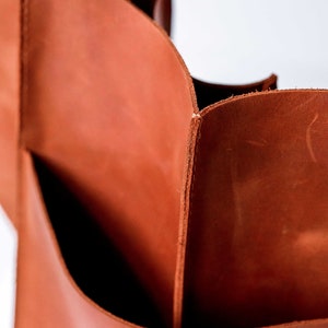 Pu&ograve; includere: Una vista ravvicinata di una borsa tote in pelle color cognac con bordi grezzi visibili e pannelli spessi e robusti. La superficie liscia e opaca della pelle mostra variazioni naturali di tono e consistenza, con pulite cuciture verticali.