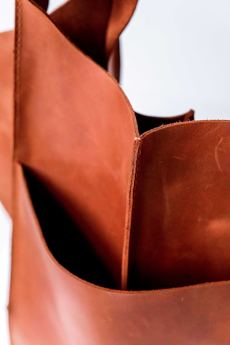 Pu&ograve; includere: Una vista ravvicinata di una borsa tote in pelle color cognac con bordi grezzi visibili e pannelli spessi e robusti. La superficie liscia e opaca della pelle mostra variazioni naturali di tono e consistenza, con pulite cuciture verticali.