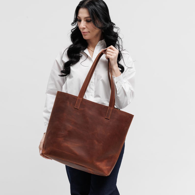 May include: A large, rectangular tote bag crafted from smooth, matte brown leather. The bag features two long, sturdy shoulder straps securely stitched to the top panel, providing a clean, structured silhouette with a spacious main compartment.
