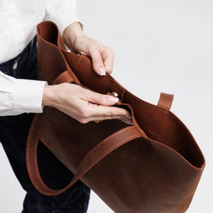 May include: A brown leather tote bag featuring a spacious open interior, two sturdy shoulder straps, and a central magnetic snap closure. The bag is constructed with visible tonal stitching along the edges and handles, showcasing a smooth finish.