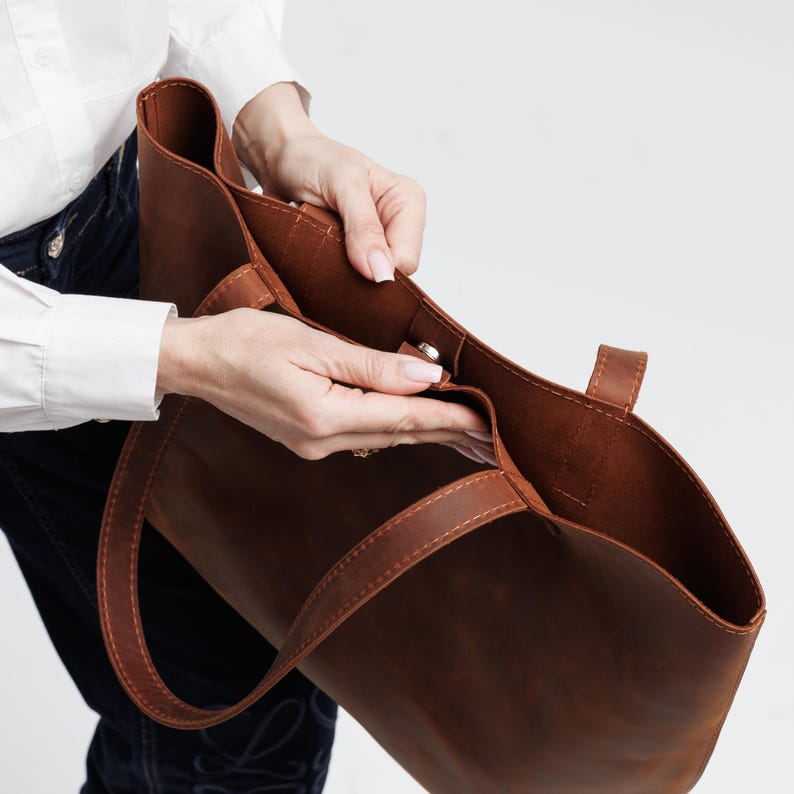 May include: A brown leather tote bag featuring a spacious open interior, two sturdy shoulder straps, and a central magnetic snap closure. The bag is constructed with visible tonal stitching along the edges and handles, showcasing a smooth finish.