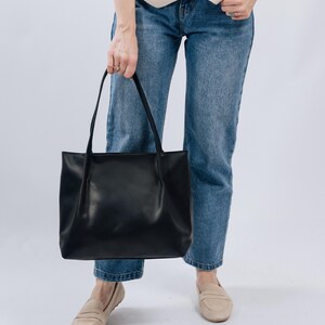 May include: A woman is wearing blue jeans and a beige vest. She is holding a black leather tote bag.