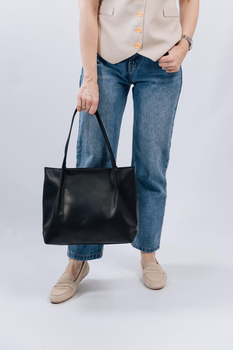 May include: A woman is wearing blue jeans and a beige vest. She is holding a black leather tote bag.