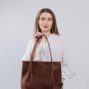 May include: A woman is holding a brown leather tote bag. The bag has a simple design with two straps and a spacious interior.