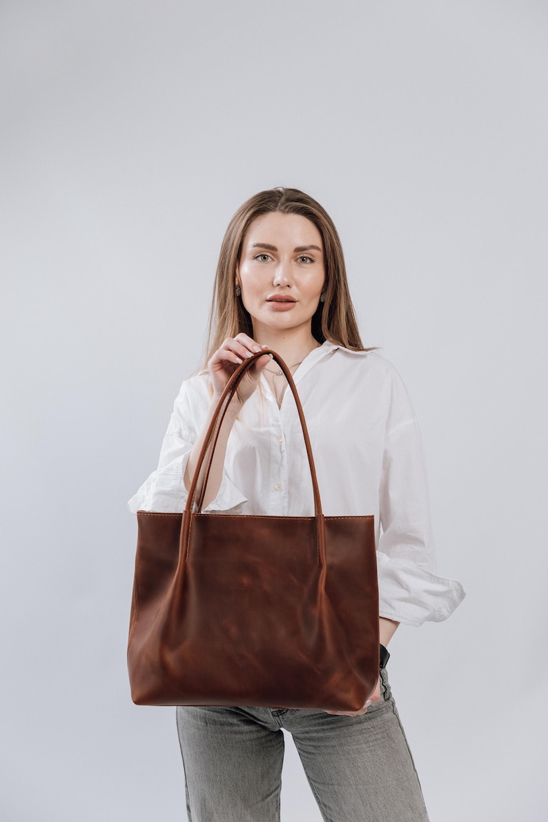 May include: A woman is holding a brown leather tote bag. The bag has a simple design with two straps and a spacious interior.