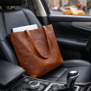 May include: A large, rectangular, cognac-brown leather tote bag with two matching shoulder straps. The bag is positioned upright on a black leather car seat, containing a thin, silver-colored laptop that is partially visible at the top opening.