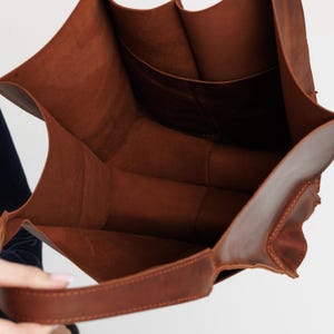 Pu&ograve; includere: Una borsa tote in pelle marrone profonda con apertura superiore e interno liscio non foderato. La borsa presenta una base ampia e robusta e un singolo manico spesso in pelle fissato sul lato, che mette in risalto la texture naturale del materiale.