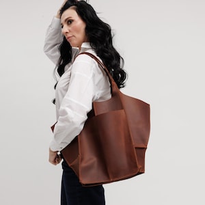 Pu&ograve; includere: Una grande borsa tote strutturata in pelle marrone con finitura opaca liscia, due ampi spallacci e una prominente tasca esterna rettangolare sul pannello frontale. La borsa presenta un design profondo e aperto con cuciture visibili sui bordi.