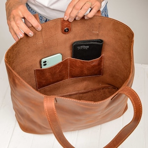 Op de afbeelding: Een bruine leren tote bag met twee binnenzakken. De tas heeft een drukknopsluiting en twee lange hengsels. Een groene telefoon en een zwarte portemonnee bevinden zich in de tas.