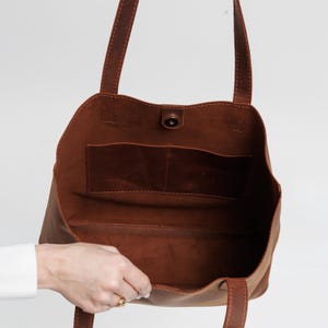 May include: A brown leather tote bag with two shoulder straps and an open top. The interior features a smooth, unlined surface with a single horizontal pocket divided into two sections. A small circular magnetic snap closure is centered at the top.