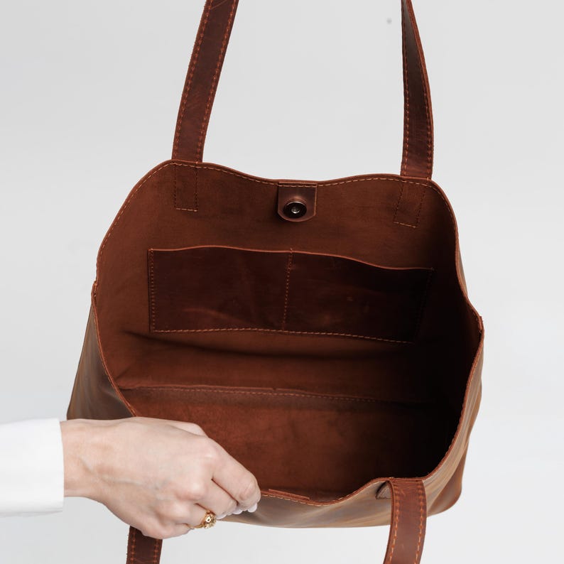 May include: A brown leather tote bag with two shoulder straps and an open top. The interior features a smooth, unlined surface with a single horizontal pocket divided into two sections. A small circular magnetic snap closure is centered at the top.