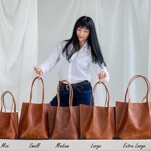 Pu&ograve; includere: Cinque borse tote in pelle marrone di diverse dimensioni allineate su una superficie bianca. Le borse sono etichettate "Mini", "Piccola", "Media", "Grande" e "Extra Grande".