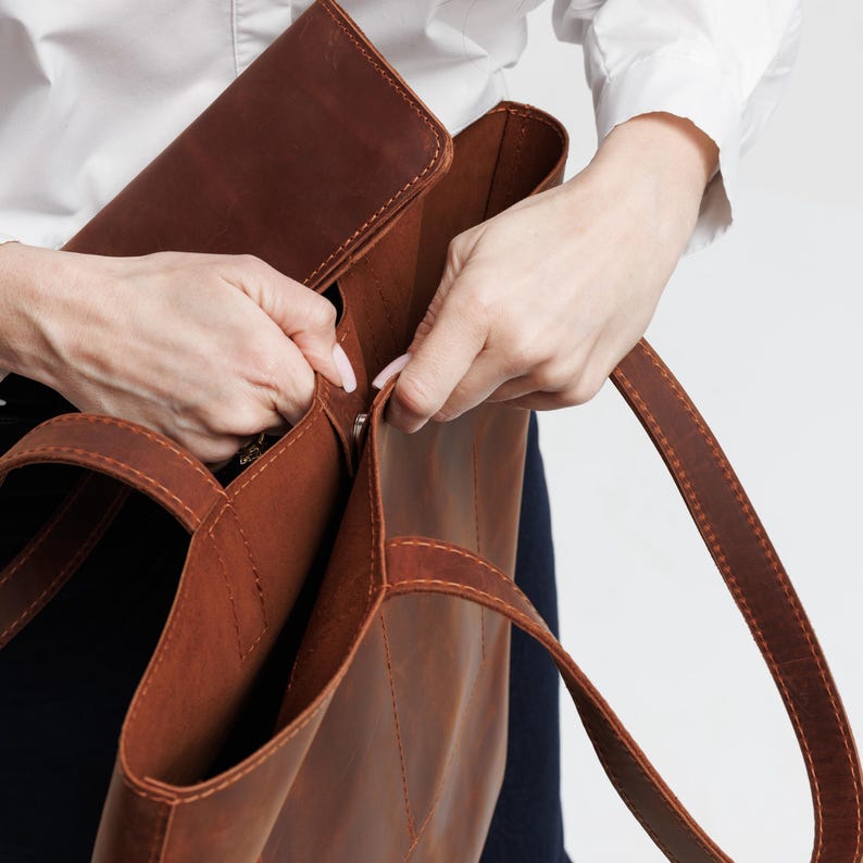May include: A large, structured brown leather tote bag featuring a smooth matte finish, reinforced stitched seams, and two sturdy shoulder straps. The bag has an open top design with a fold-over flap closure secured by a metal snap button fastener.