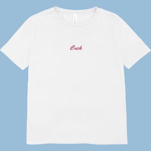 Puede incluir: Camiseta blanca de cuello redondo con la palabra "Cuck" bordada en rosa cursiva en el pecho. La camiseta está hecha de un material suave y tiene mangas cortas. El fondo es de color azul claro liso.