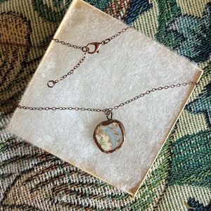May include: A necklace featuring a small, irregular pendant made from a piece of vintage ceramic with a light blue and white floral pattern, encased in a copper-colored metal frame and attached to a matching copper-colored metal chain.